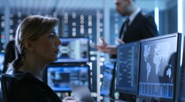 Female Government Employee Works In A Monitoring Room. In The Background Supervisor Holds Briefing. Possibly Government Agency Conducts Investigation.